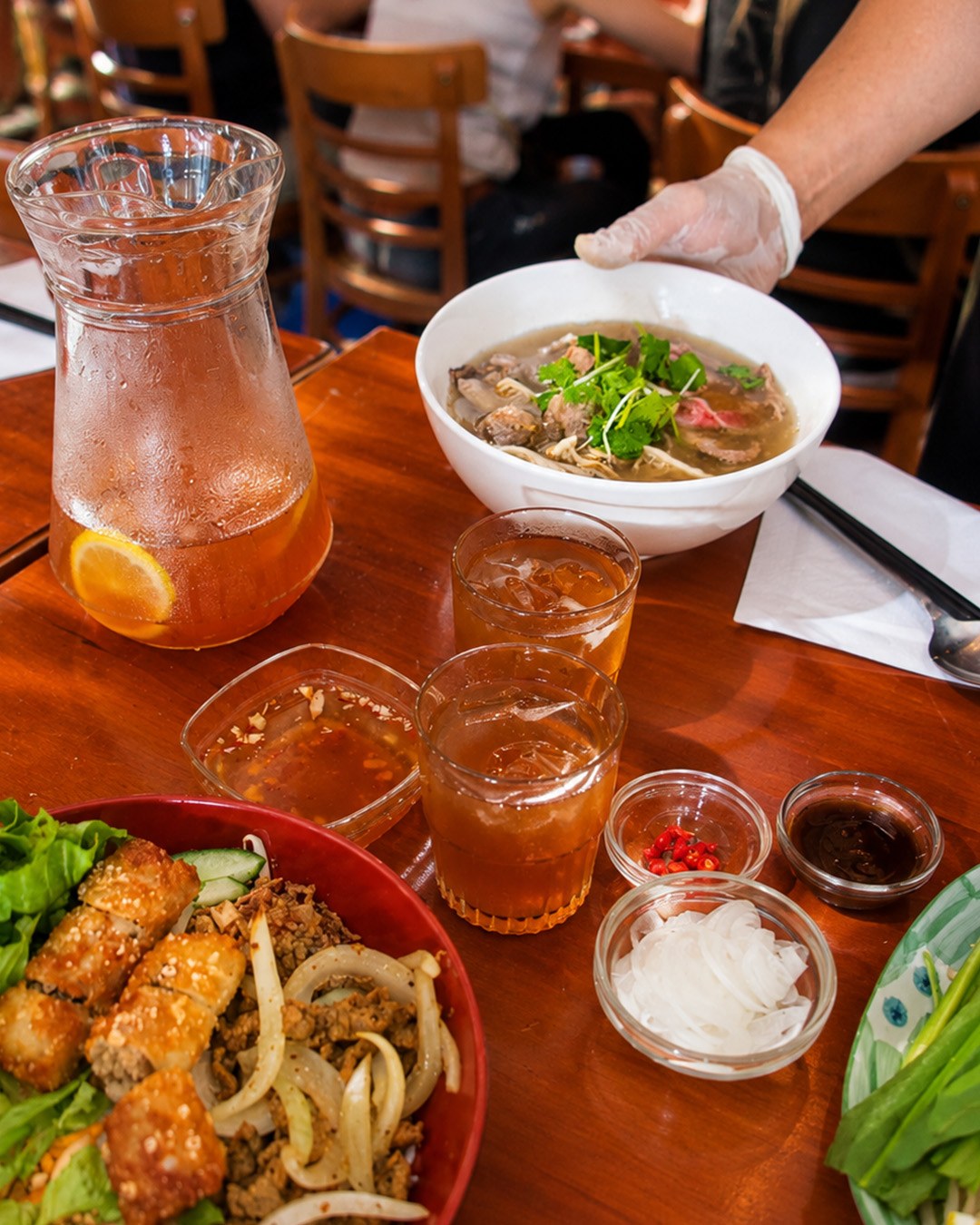 Une table servie avec un bol de pho, un plat garni, des condiments et une carafe de thé glacé